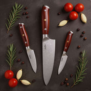 Set of three knives with red handles on a dark surface with vegetables and herbs.
