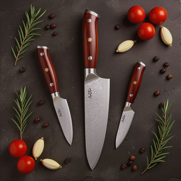 Set of three knives with red handles on a dark surface with vegetables and herbs.