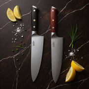 Two chef's knives with different handles on a dark marble surface with lemon slices and herbs.